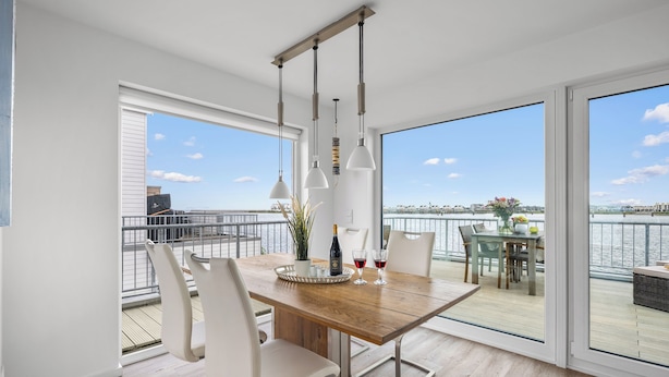 Un comedor moderno con una mesa de madera, sillas blancas y luces colgantes. Cuenta con ventanas amplias con vista al agua y al paisaje urbano.
