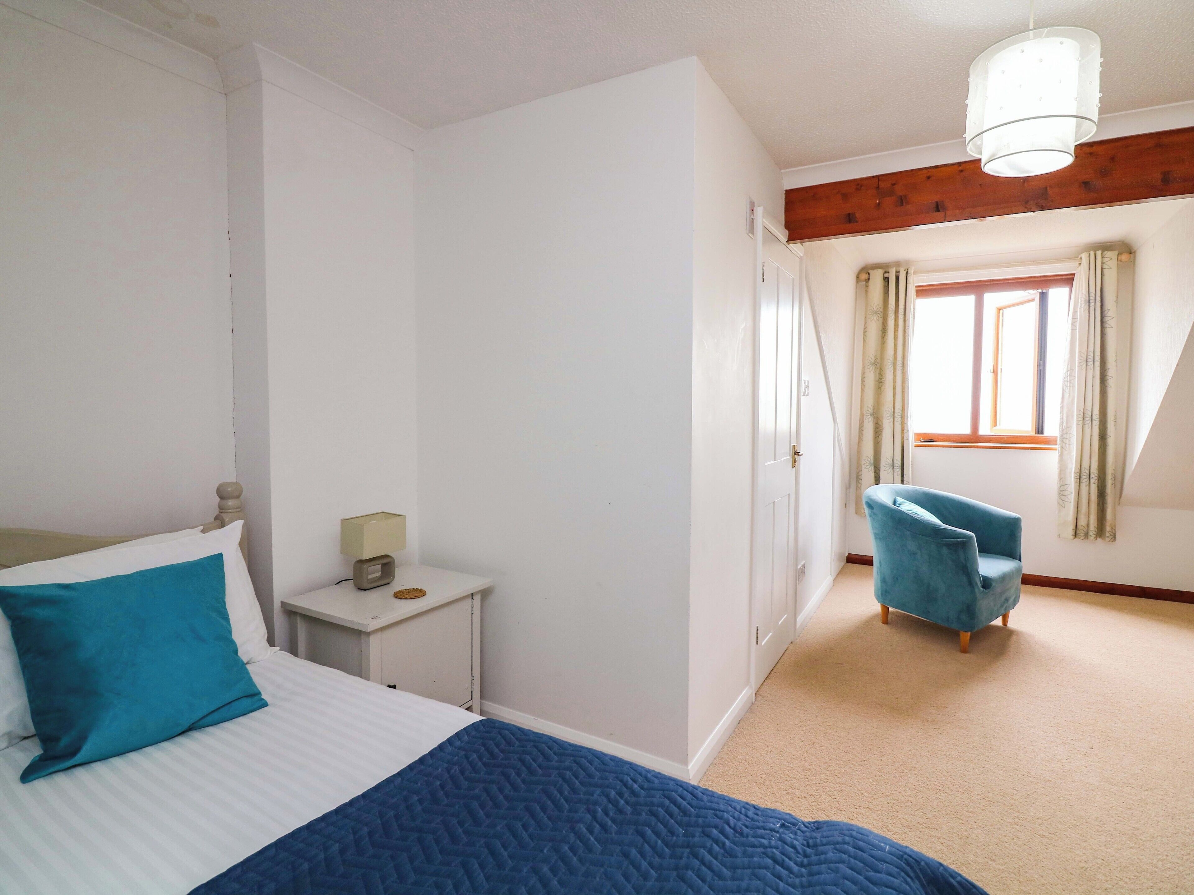 A bedroom with a bed, a nightstand, a chair, a window with curtains, and a wooden ceiling beam.