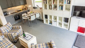 A modern kitchen with grey cabinets, a dining area with a white table and chairs, a living area with a sofa and a coffee table, and a bookshelf with various items.
