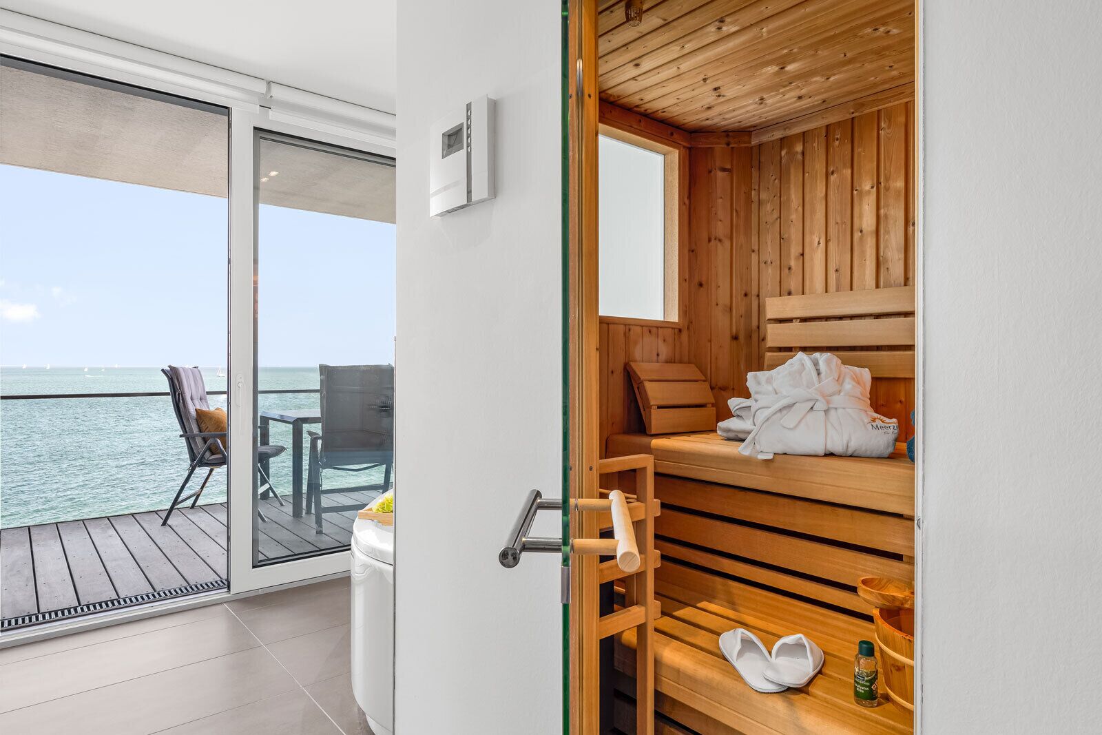 Une chambre moderne dotée d’un sauna en bois, d’une porte en verre donnant sur un balcon avec vue sur la mer, et d’une petite table avec une chaise.