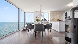 Uma cozinha moderna com fogão embutido, área de jantar com mesa redonda e cadeiras, e grandes janelas com vista para o mar.