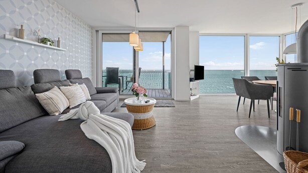Uma sala de estar moderna com um sofá cinza, uma mesa de centro de vime redonda e uma TV sobre um armário cinza. Uma grande janela oferece vista para o mar.