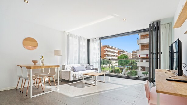 A modern living room with a dining area, a sofa, a TV, and a large window with a view of buildings. - Appartamento di fronte al mare, piscina, aria condizionata, parcheggio ascensore (El Mas Mel)