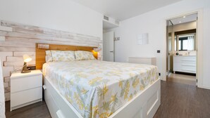A bedroom with a wooden headboard, a bedside table with a lamp, a bed with a floral bedspread, a walk-in closet, and a bathroom with a mirror. - Appartamento di fronte al mare, piscina, aria condizionata, parcheggio ascensore (El Mas Mel)
