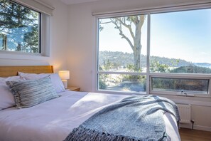 A bedroom with a large window, a bed with white bedding, and a view of a forested area. - Sjøro • Stunning Sea Views • Beach Access • Sauna (Kingston Beach)