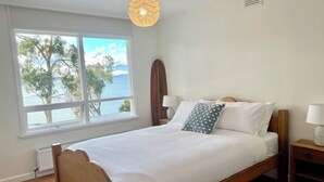 A bedroom with a wooden bed, white bedding, a view of the sea, and a window with a view of trees and mountains. - Beach house with sauna. Stunning views, cosy renovated mid-century gem (Kingston Beach)