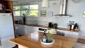 A modern kitchen with a central island, bar stools, a hanging light fixture, and a view of the outdoors through large windows. - Beach house with sauna. Stunning views, cosy renovated mid-century gem (Kingston Beach)