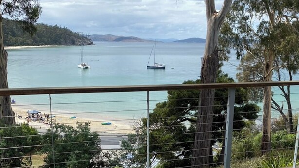 A balcony with wicker furniture, a potted plant, and a view of a beach and sailboats. - Beach house with sauna. Stunning views, cosy renovated mid-century gem (Kingston Beach)