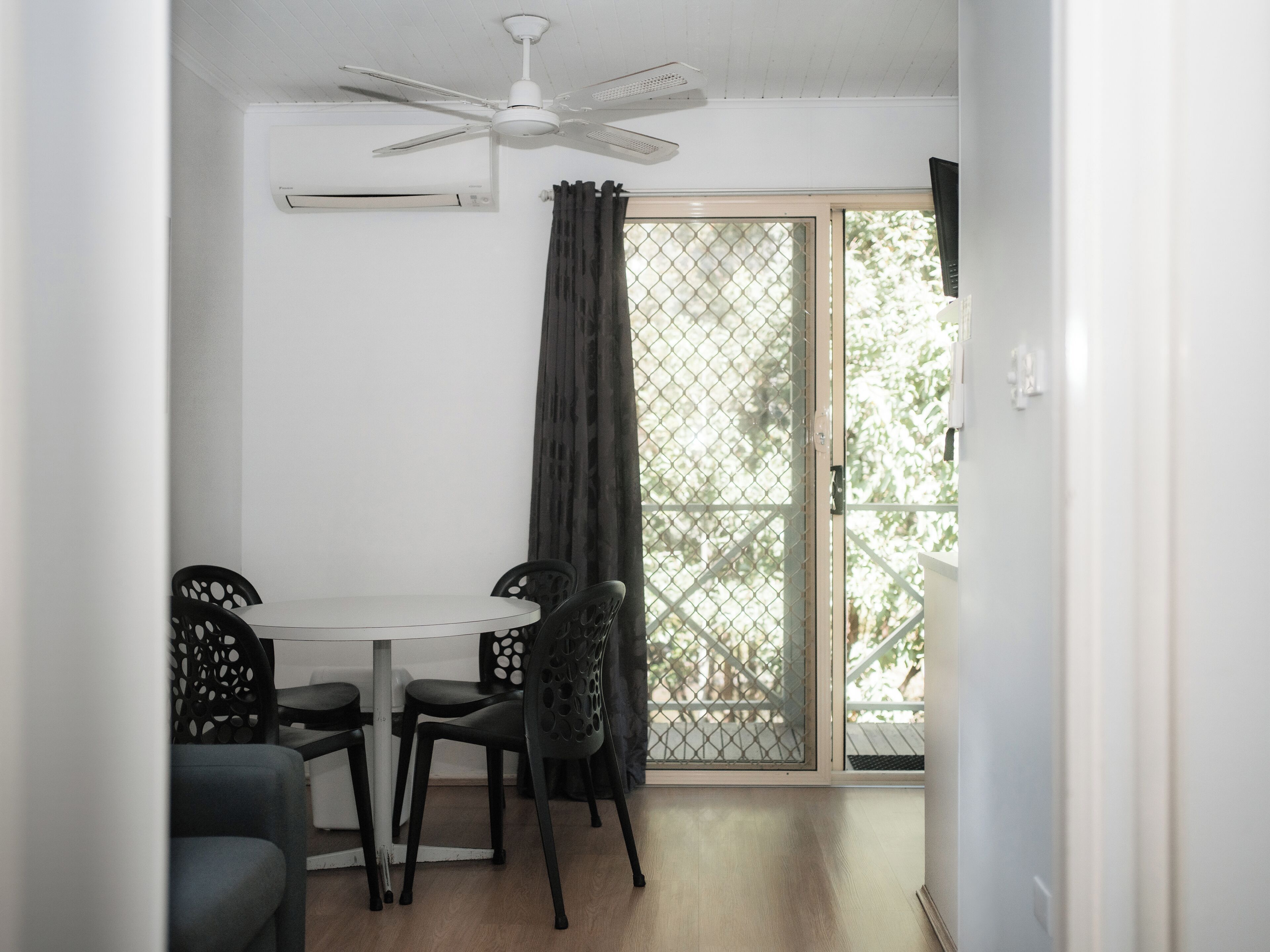 A room with a ceiling fan, a window with a metal screen, a white table with four chairs, and a wooden floor.