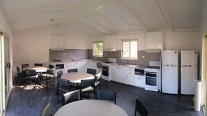 A modern kitchen with white cabinets, a dining area with round tables and chairs, and a large window providing natural light. - Warburton Holiday Park (Warburton)