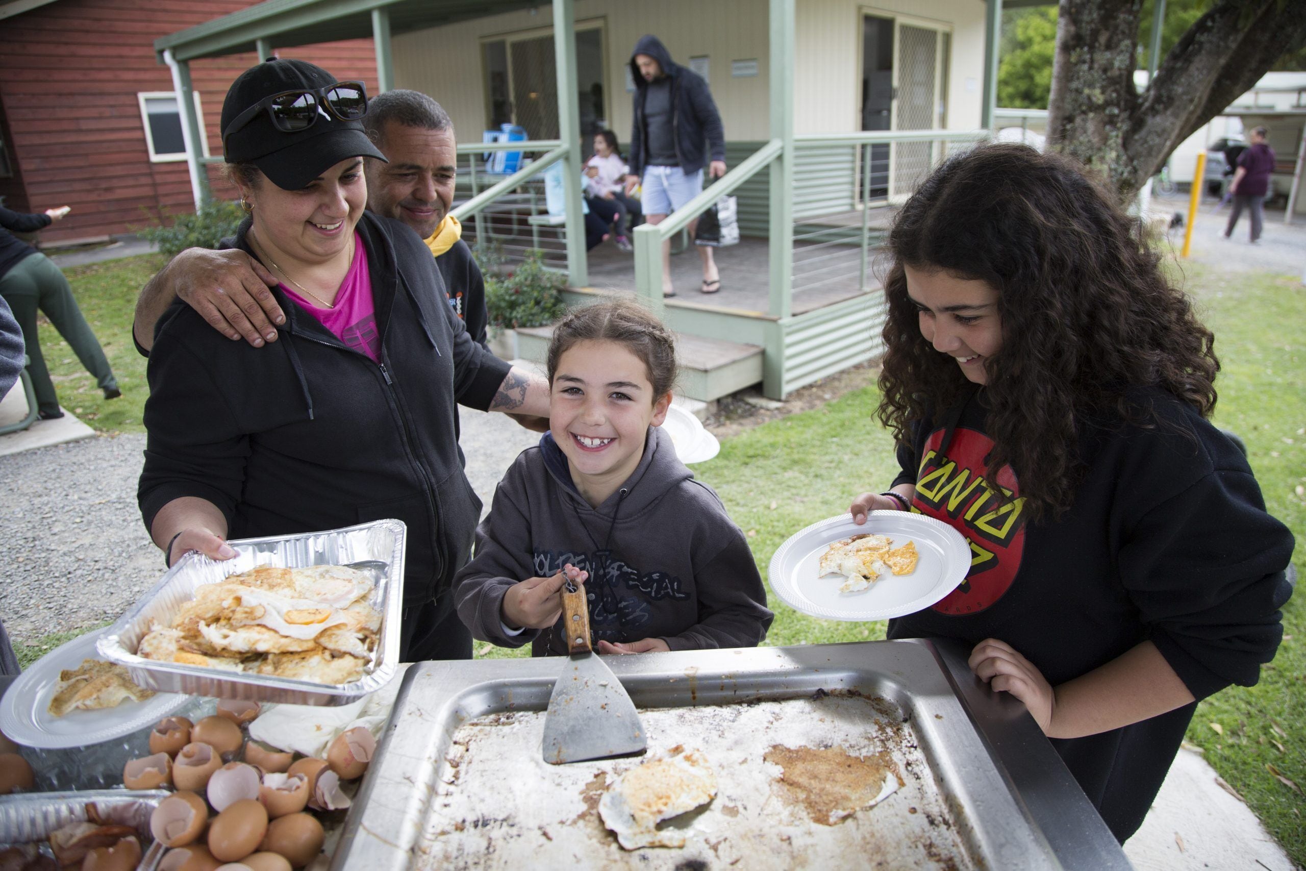 A group of people gathered around a food serving area with trays of food and a spatula.