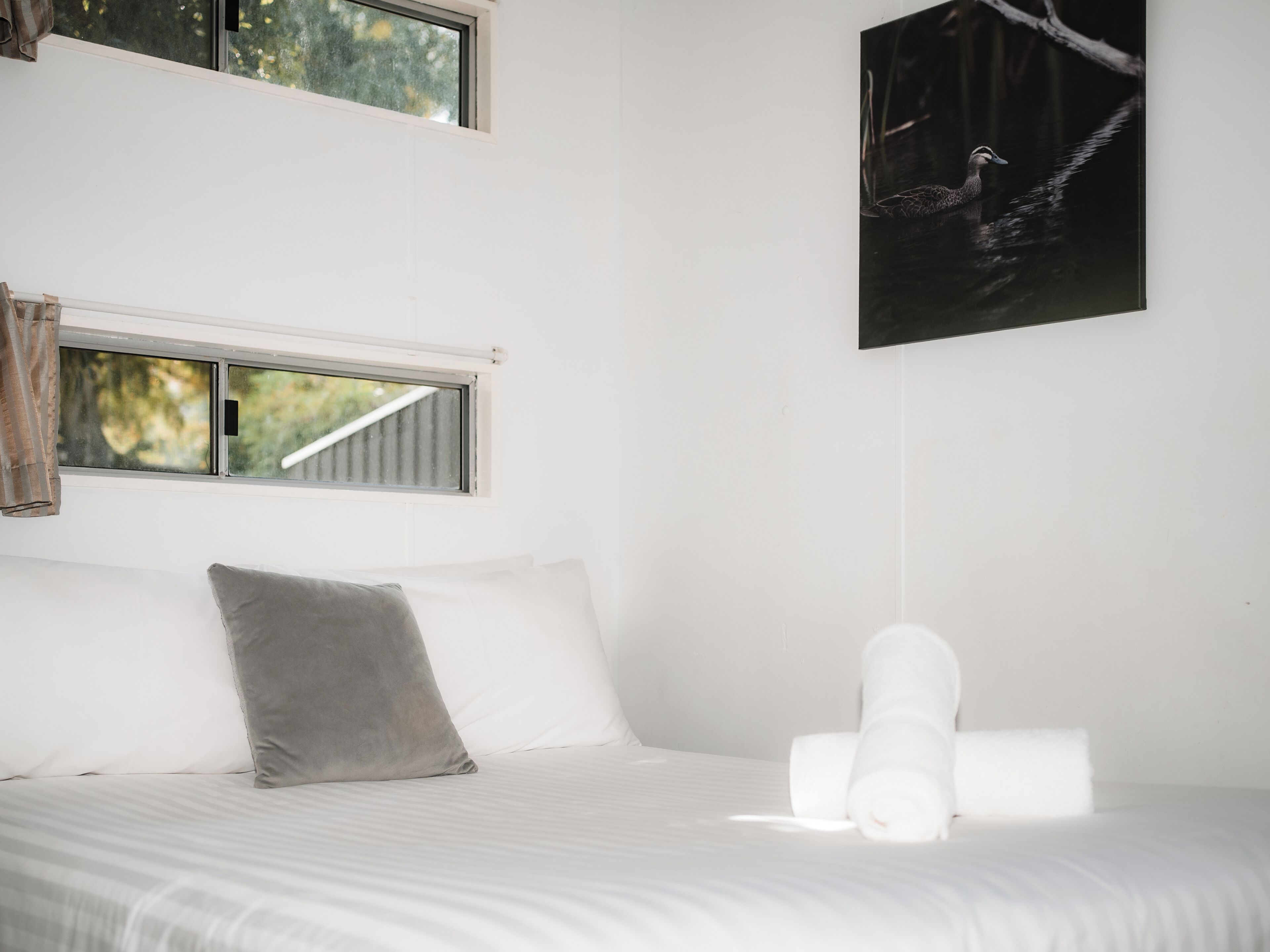 Ein Bett mit weißen Laken, einem grauen Kissen und einem aufgerollten weißen Handtuch.