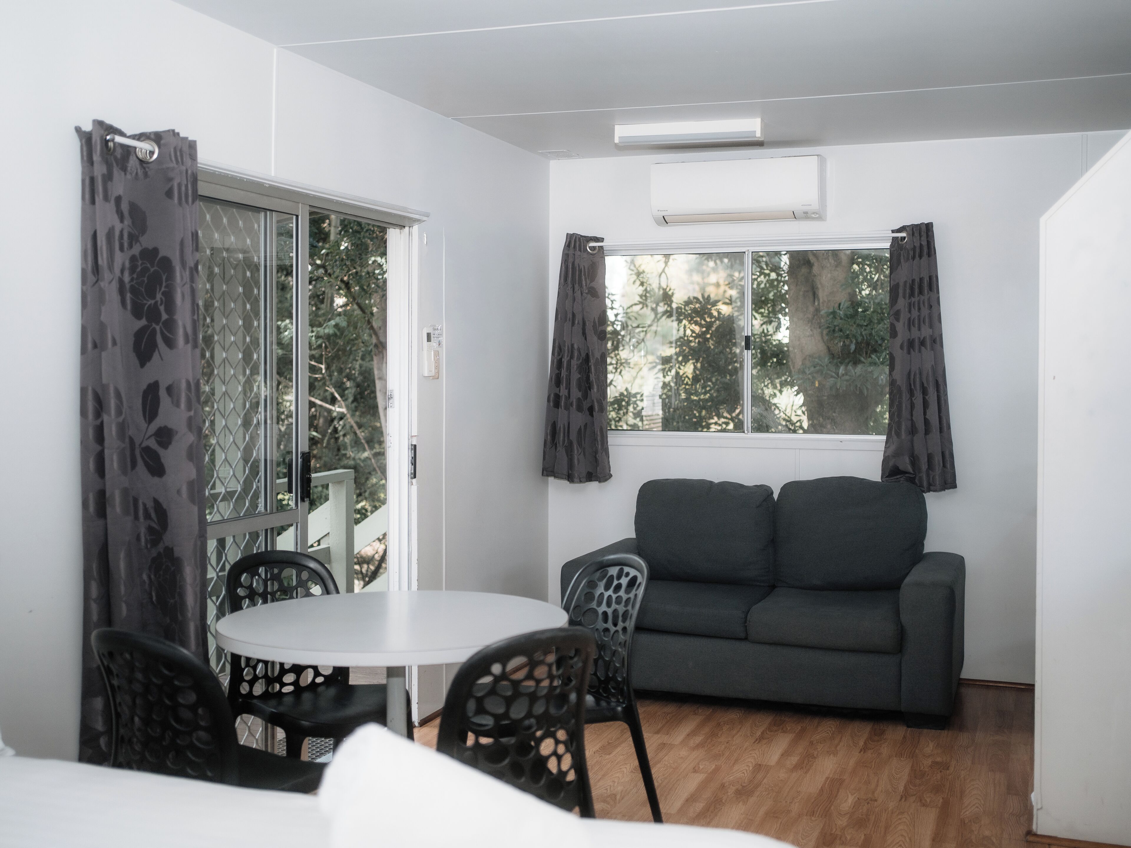 A room with a sofa, a dining table with chairs, a window with curtains, and an air conditioning unit.