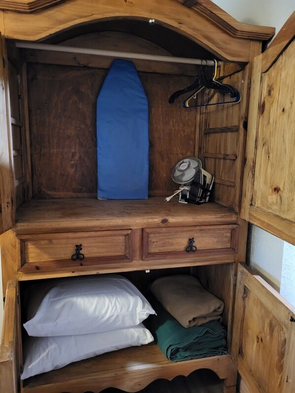 A wooden wardrobe with a blue ironing board, a fan, and folded clothes.