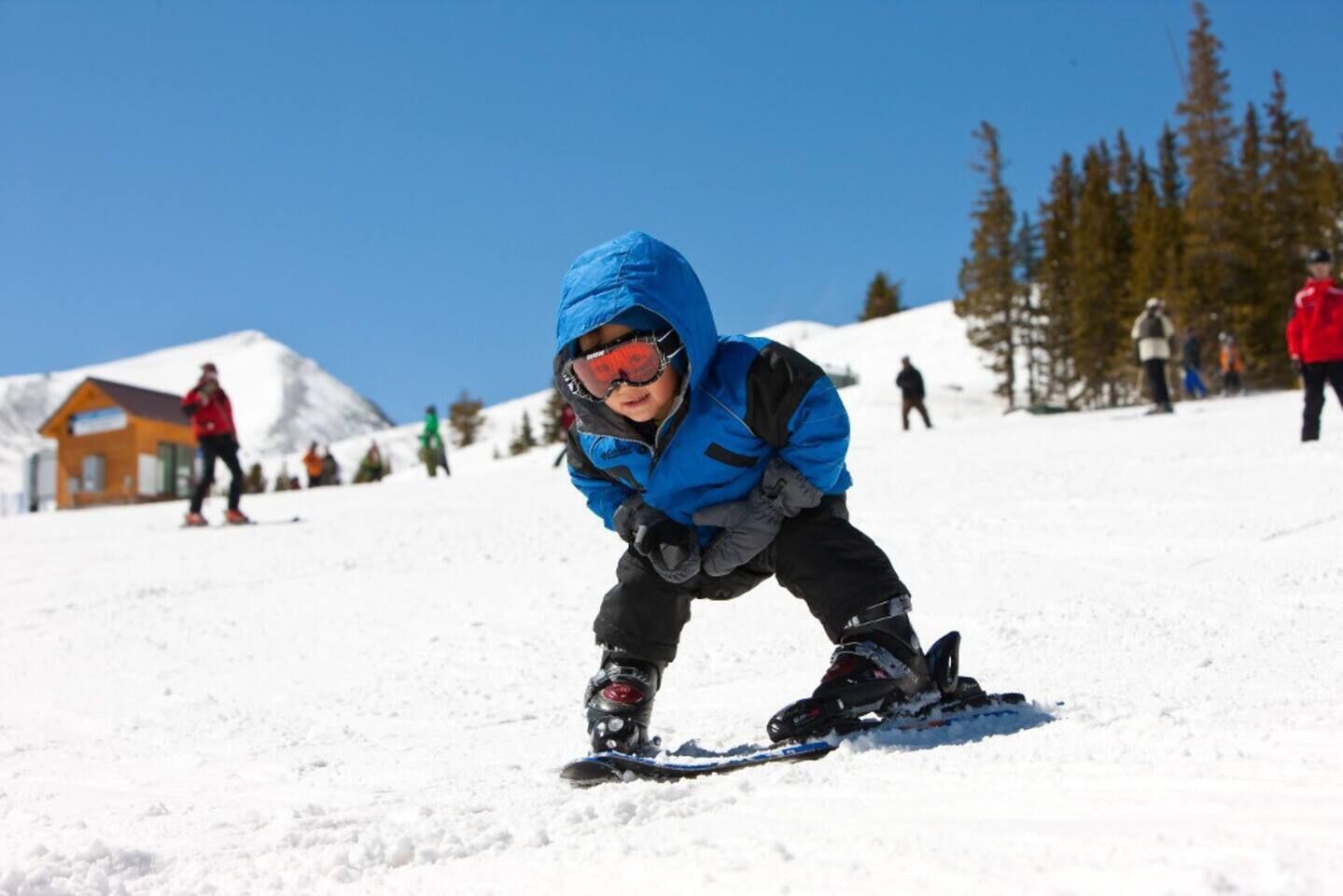 Un enfant fait de la planche à neige sur une pente enneigée, sous un ciel bleu dégagé.