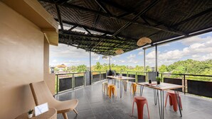 Uma área de estar coberta ao ar livre, com mesa e cadeiras, uma cadeira de vime e vista para áreas verdes e edifícios ao fundo.
