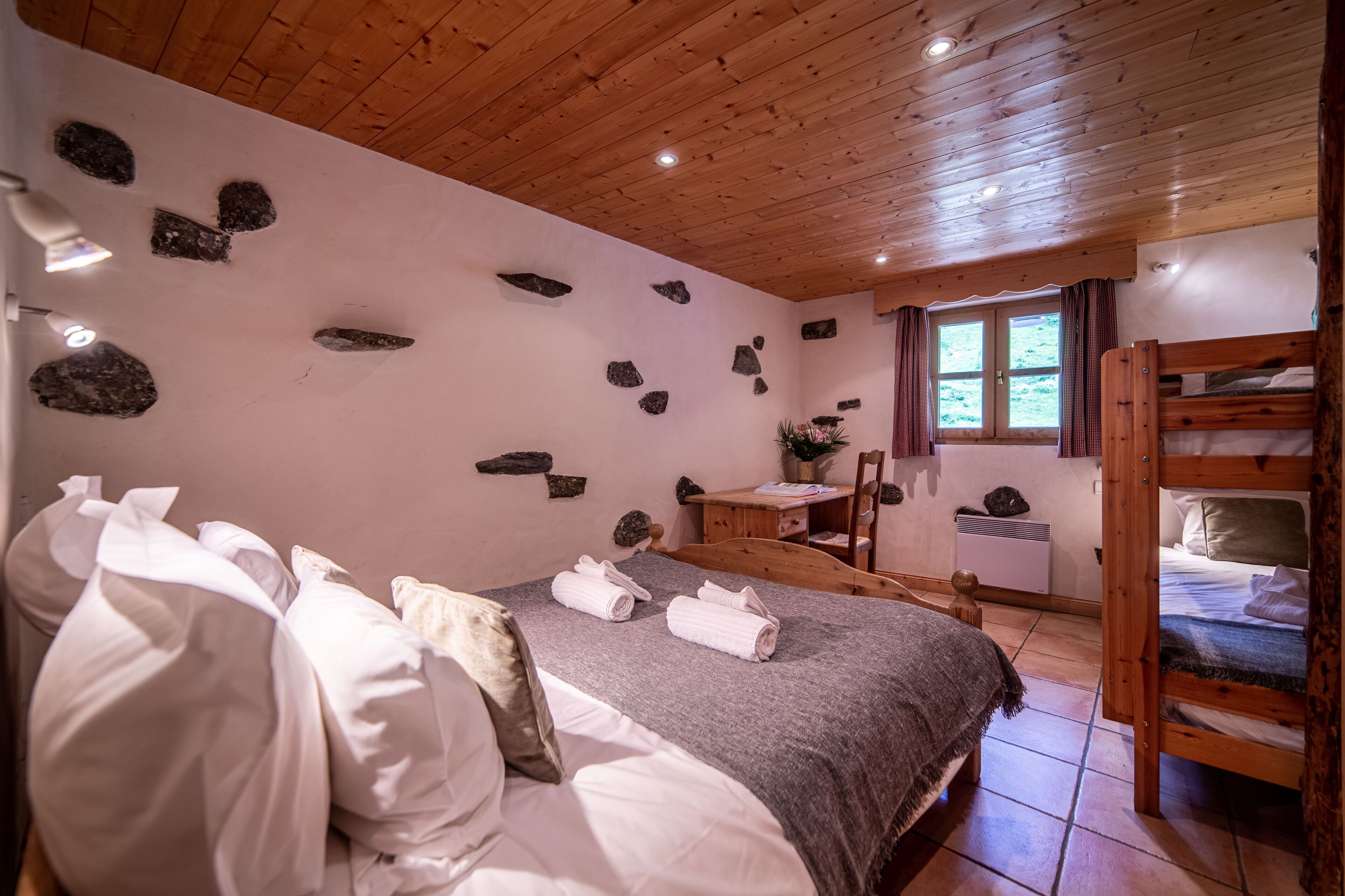 A cozy bedroom with a wooden ceiling, a bunk bed, a desk with a chair, and a window with curtains.