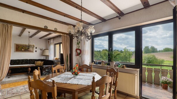 A dining area with a table set for four, surrounded by wooden chairs with green cushions. A chandelier hangs from the ceiling, and a large window offers a view of greenery outside.