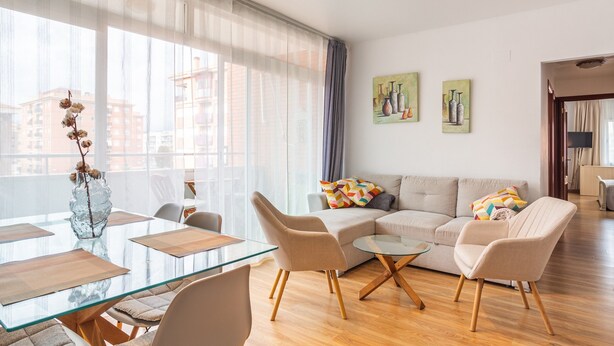 A modern living room with a glass dining table, beige sofas, and a view of buildings through large windows.