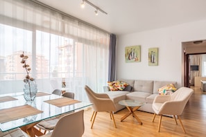 Een moderne woonkamer met een glazen eettafel, beige banken en een uitzicht op gebouwen door grote ramen.