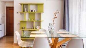 A modern dining area with a glass table, white chairs, a green bookshelf, and a vase with flowers.