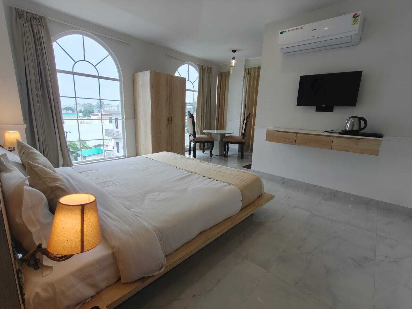 A hotel room with a large bed, a TV mounted on the wall, a small table with chairs, and an air conditioning unit.