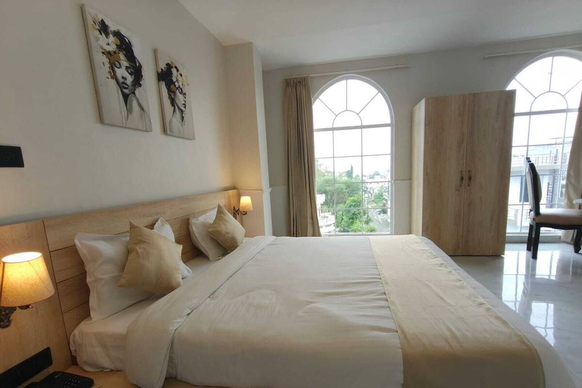 A hotel room with a large bed, two wall-mounted lamps, a wardrobe, and a view of the city through arched windows.
