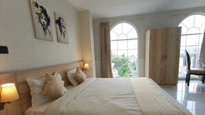 Habitación de hotel con una cama grande, dos lámparas de pared, un armario y vistas a la ciudad a través de ventanales arqueados.