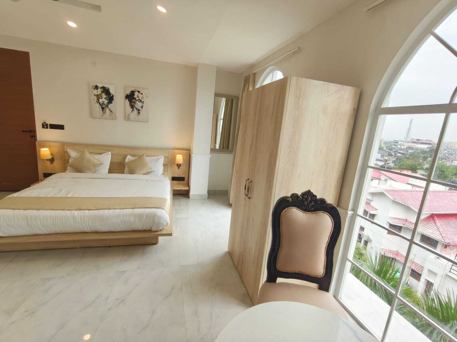 A bedroom with a large bed, a wardrobe, a chair, and a view of buildings through a window.