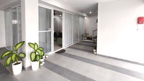 Un pasillo amplio con diseño moderno, con un ventanal corredero que da a una habitación, plantas en macetas y un detector de incendios rojo en la pared.