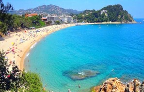 A coastal beach with clear blue waters, a sandy shore, and buildings in the background.
