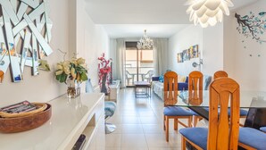 A modern living room with a glass dining table, wooden chairs, a chandelier, and a view to the dining area. - Apartment 'Verano Go2lloret' with Mountain View, Wi-Fi and Air Conditioning (Lloret de Mar)