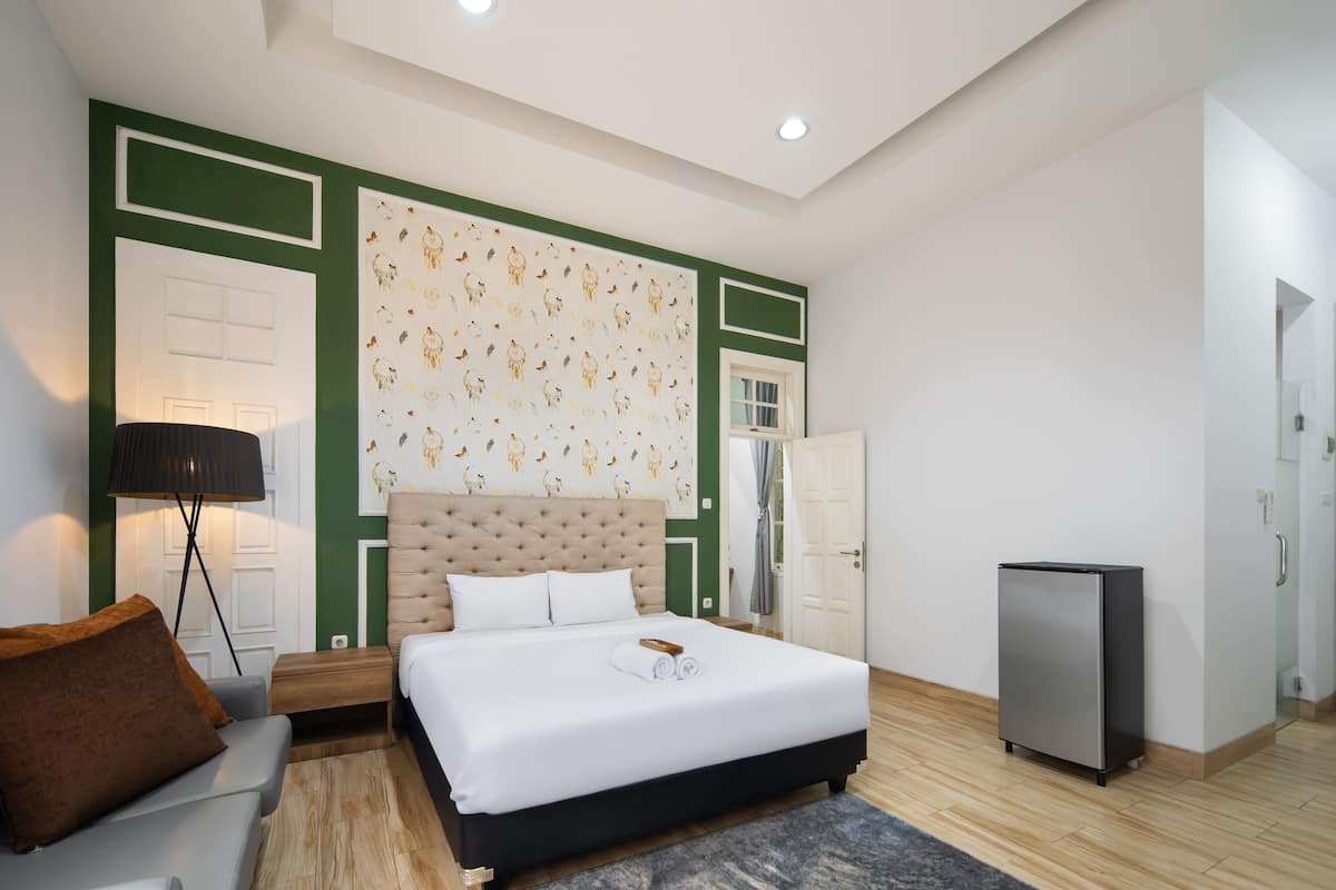 A modern hotel room with a large bed, a bedside lamp, a small table, and a refrigerator.