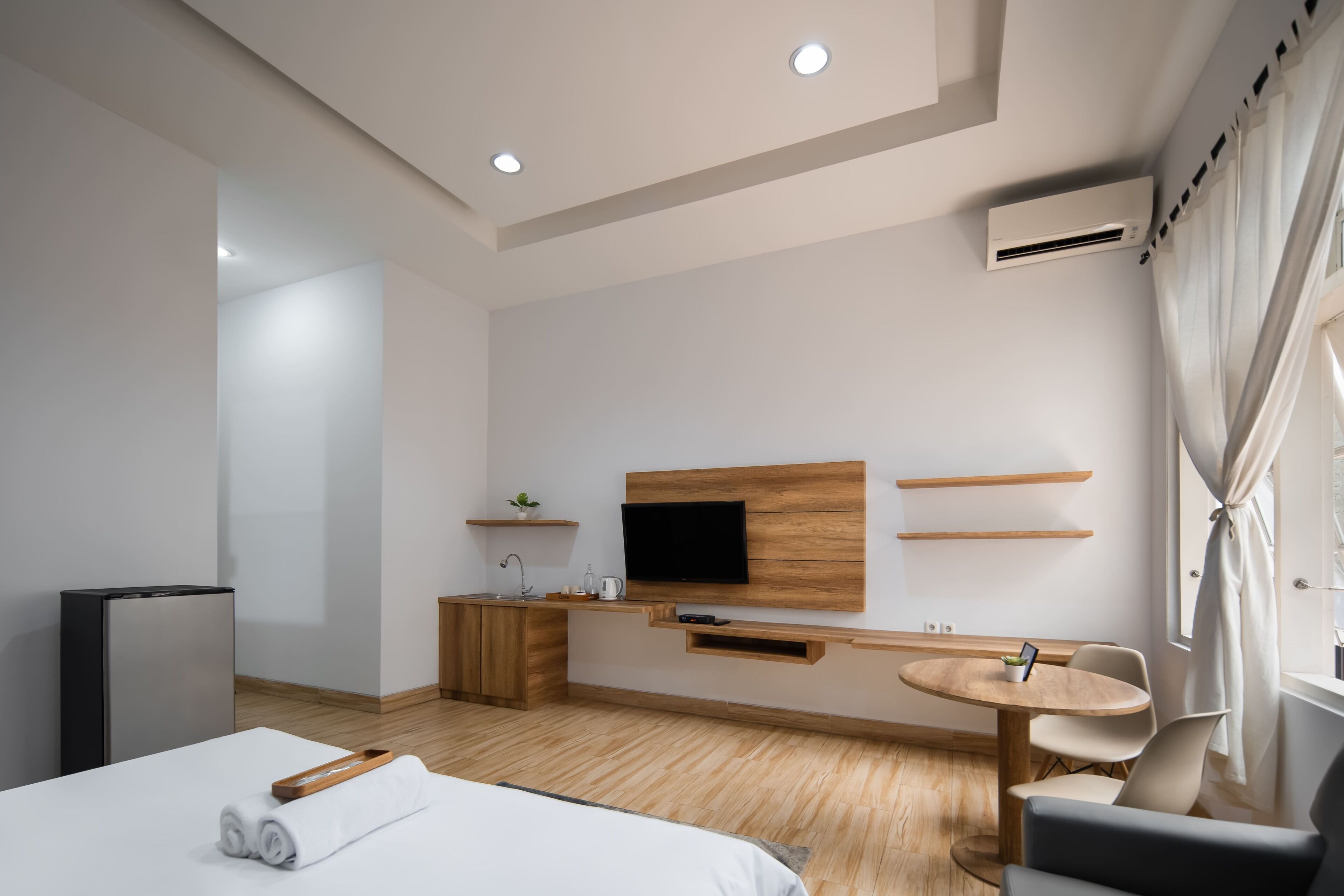 A modern hotel room with a bed, a small kitchenette, a TV mounted on the wall, and a window with curtains.