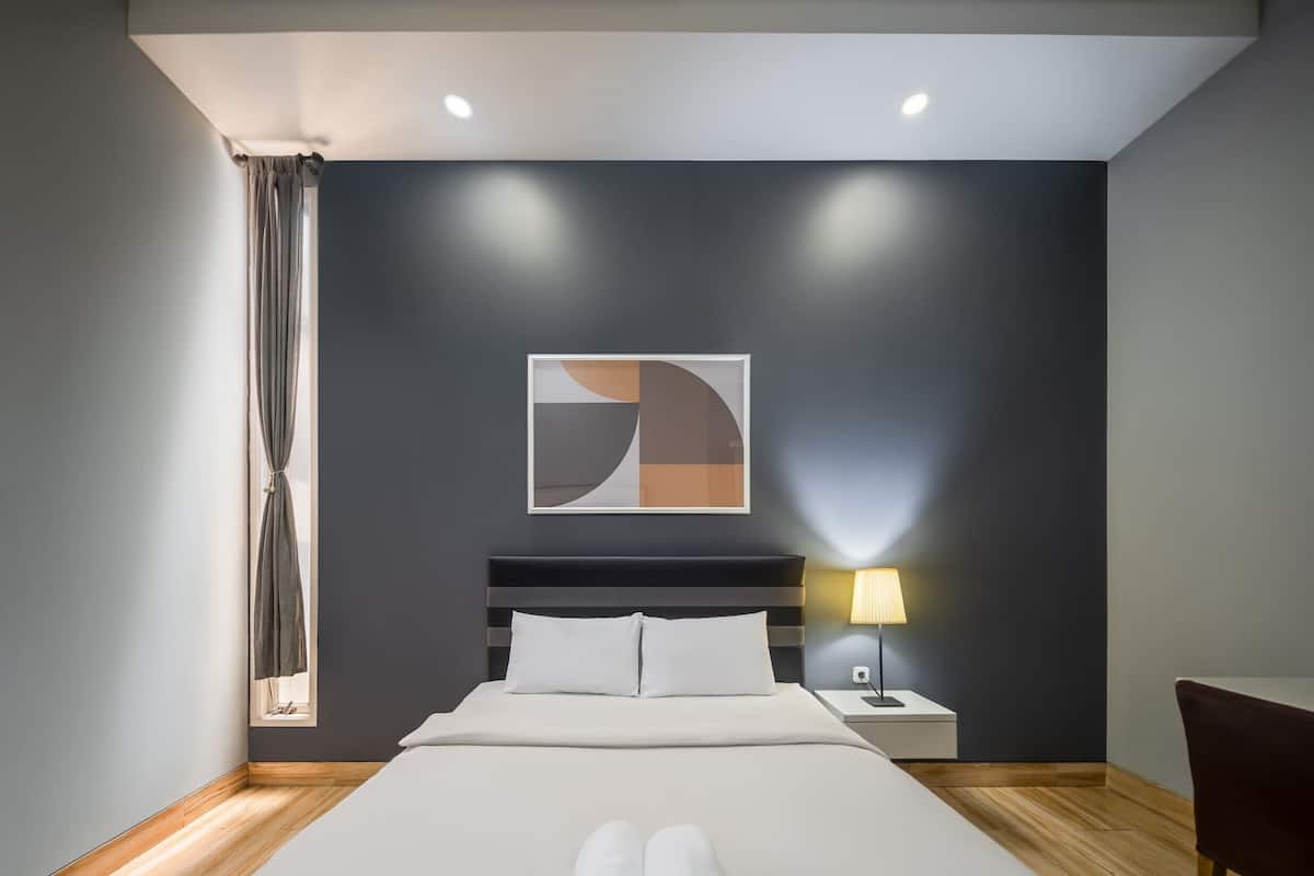 A modern hotel room with a large bed, a nightstand with a lamp, a window with curtains, and a wall art piece.