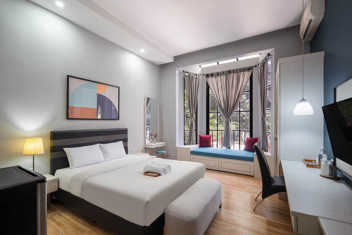 A modern hotel room with a large bed, a desk, a chair, a window with curtains, a small table with a lamp, and a painting on the wall.