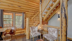 Interior de una cabaña de madera con escalera, paredes de madera, zona de comedor con sillas y una mesa.