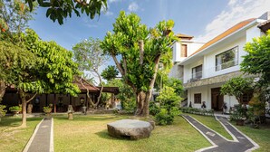 A courtyard with a paved walkway, a stone bench, and a large tree. - Cove Puri Maharani (Denpasar)