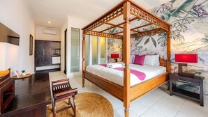 A four-poster bed with a wooden frame, a bedside table with a lamp, a desk with a chair, and a woven mat. - Cove Puri Maharani (Denpasar)