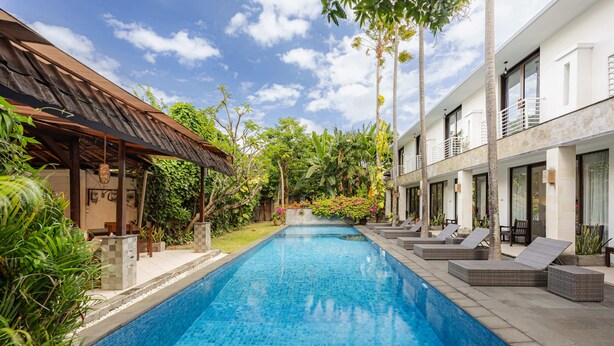 A swimming pool with lounge chairs and a covered area, surrounded by lush greenery and a two-story building. - Cove Puri Maharani (Denpasar)