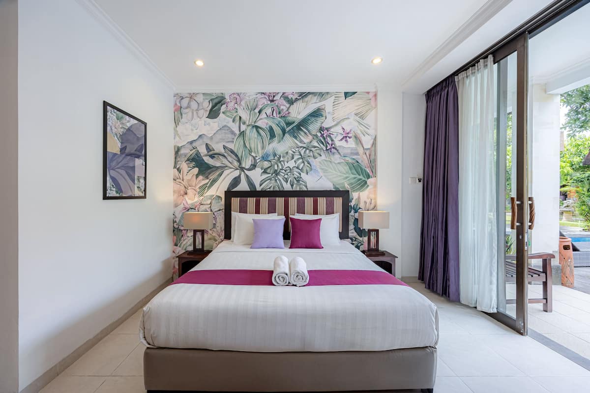 A hotel room with a large bed, two bedside lamps, a tropical-themed wall mural, and a sliding glass door to the outside.