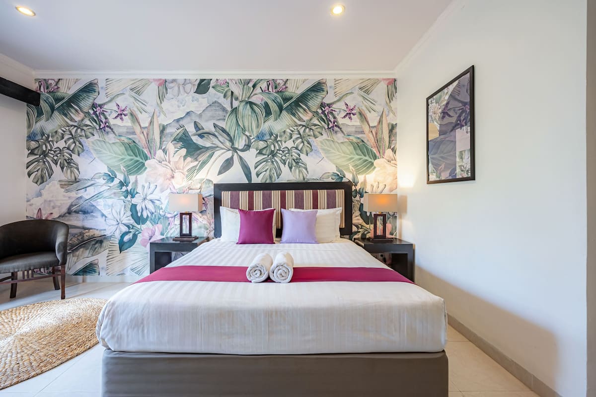 A hotel room with a large bed, a headboard, a nightstand, a lamp, a chair, and a tropical-themed wall mural.
