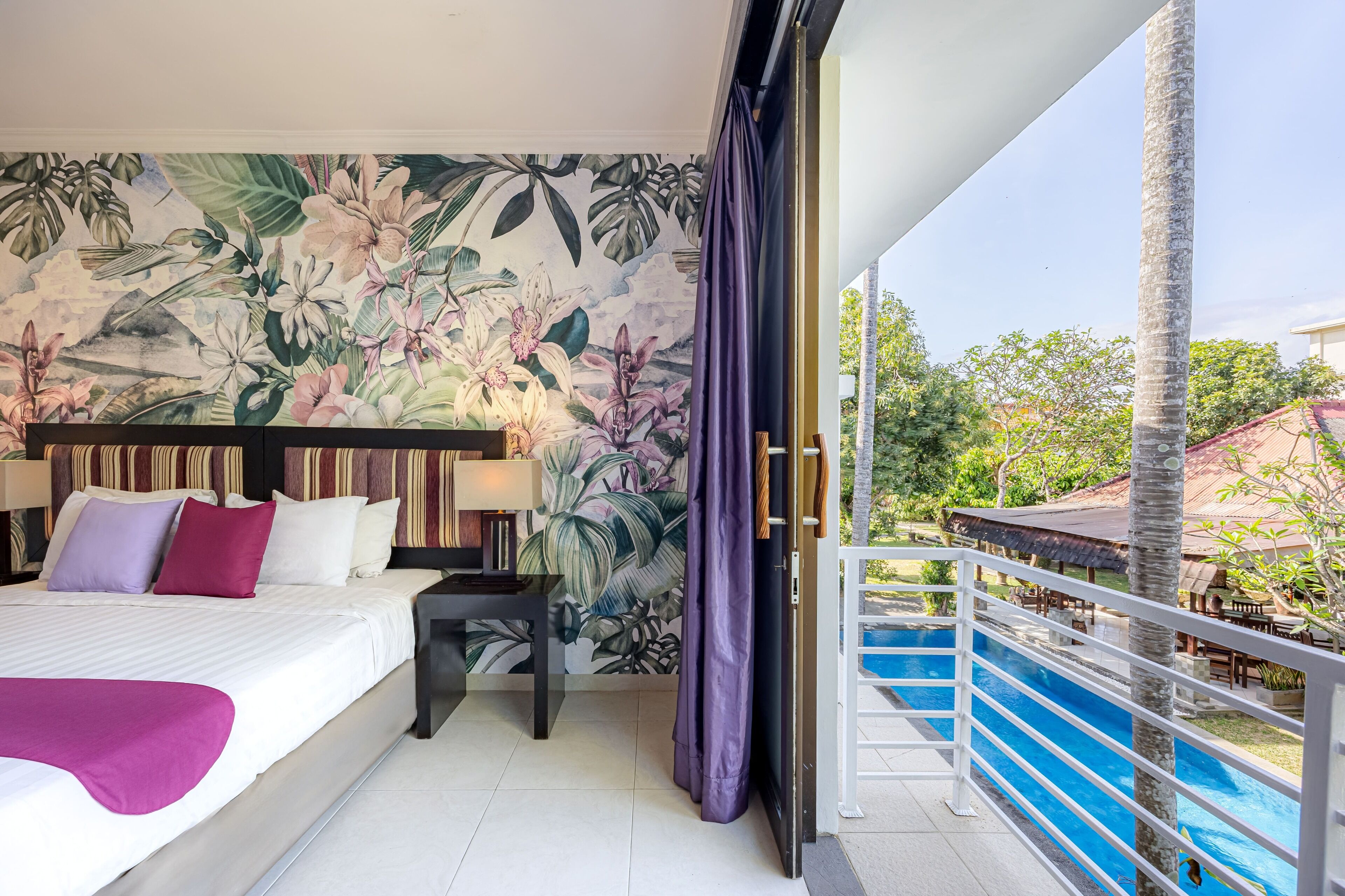 Une chambre d’hôtel avec un lit, une table de chevet, une lampe, une fresque murale aux motifs tropicaux et un balcon donnant sur une piscine.