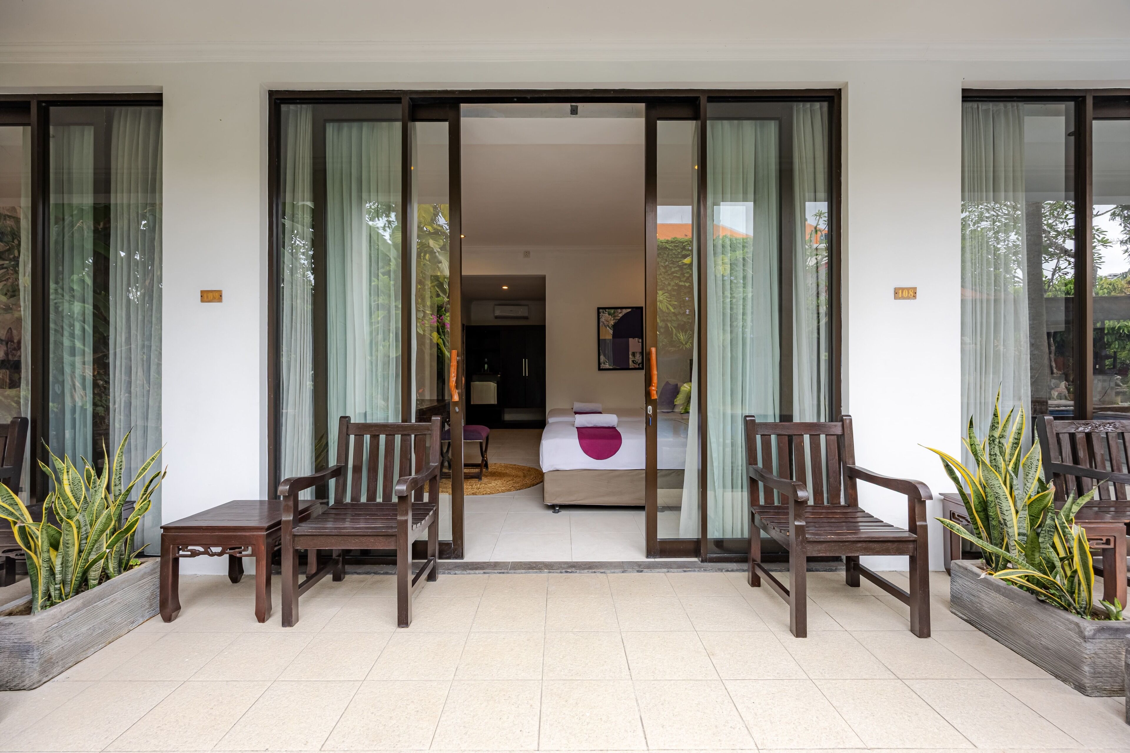 Entrada de hotel con puertas de vidrio correderas, una cama con un almohadón rosa, y sillas y mesas de madera.