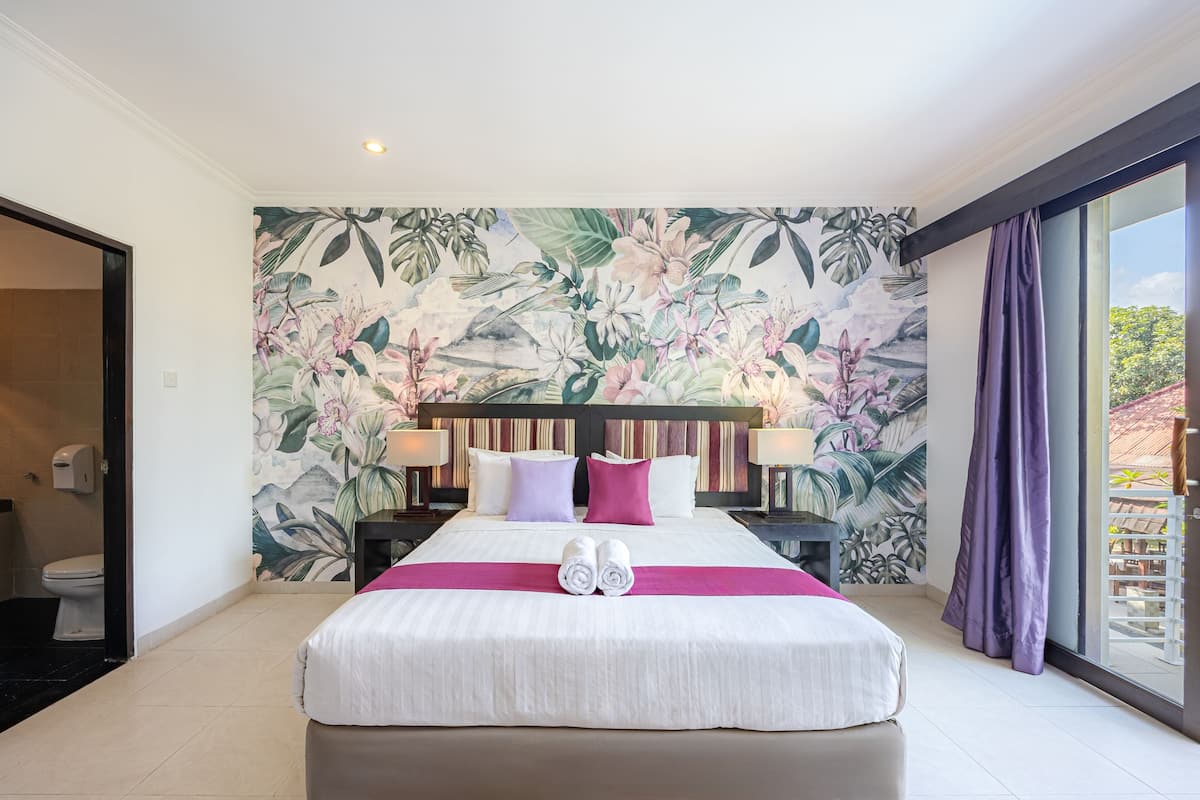 A hotel room with a large bed, a tropical floral mural, a balcony with purple curtains, and a view of greenery.
