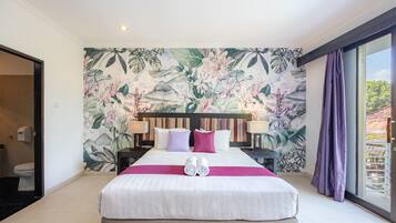 Ein Hotelzimmer mit einem großen Bett, einer tropischen Blumenmalerei, einem Balkon mit violetten Vorhängen und Blick ins Grüne.