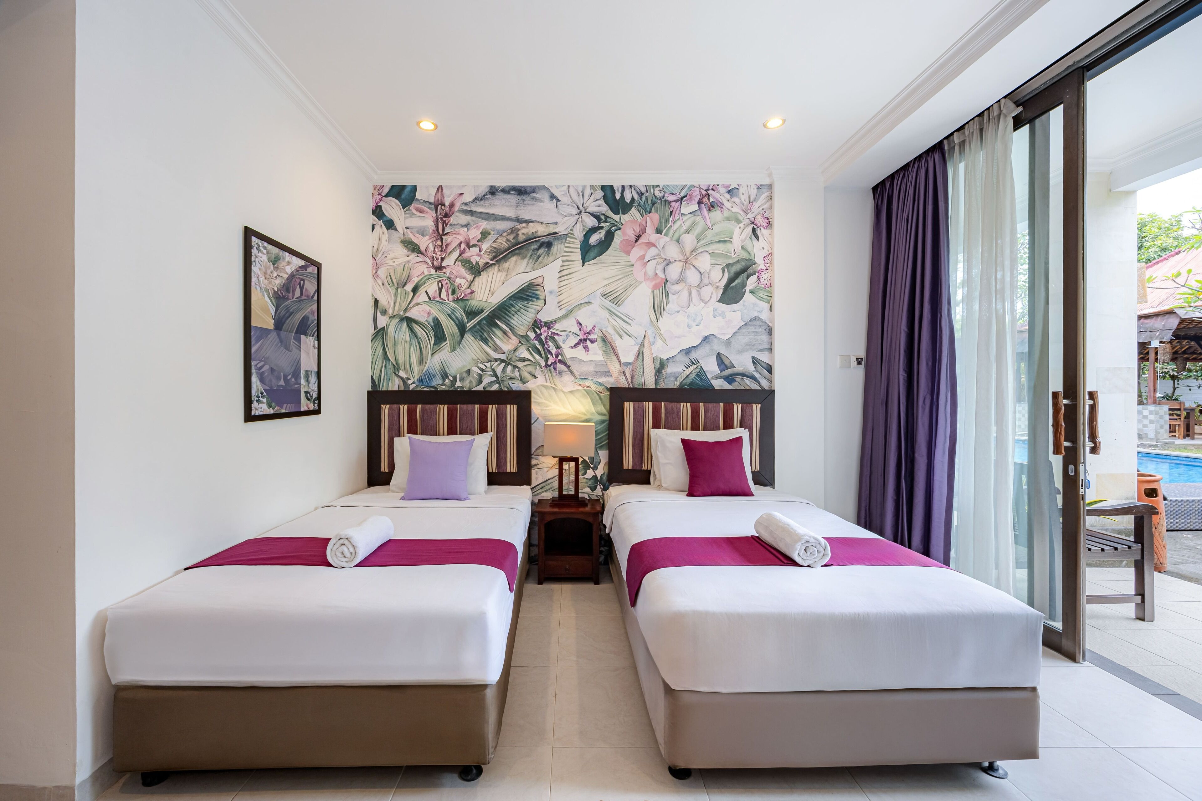 Une chambre d’hôtel avec deux lits, une grande fresque florale sur le mur, une petite table avec une lampe et une vue sur l’extérieur.