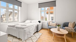 A modern bedroom with a bed, two chairs, a small table, and large windows offering a view of buildings.