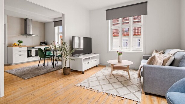 A modern living room with a grey sofa, a coffee table, and a TV. There is a dining area with a table and chairs, and a kitchen in the background.