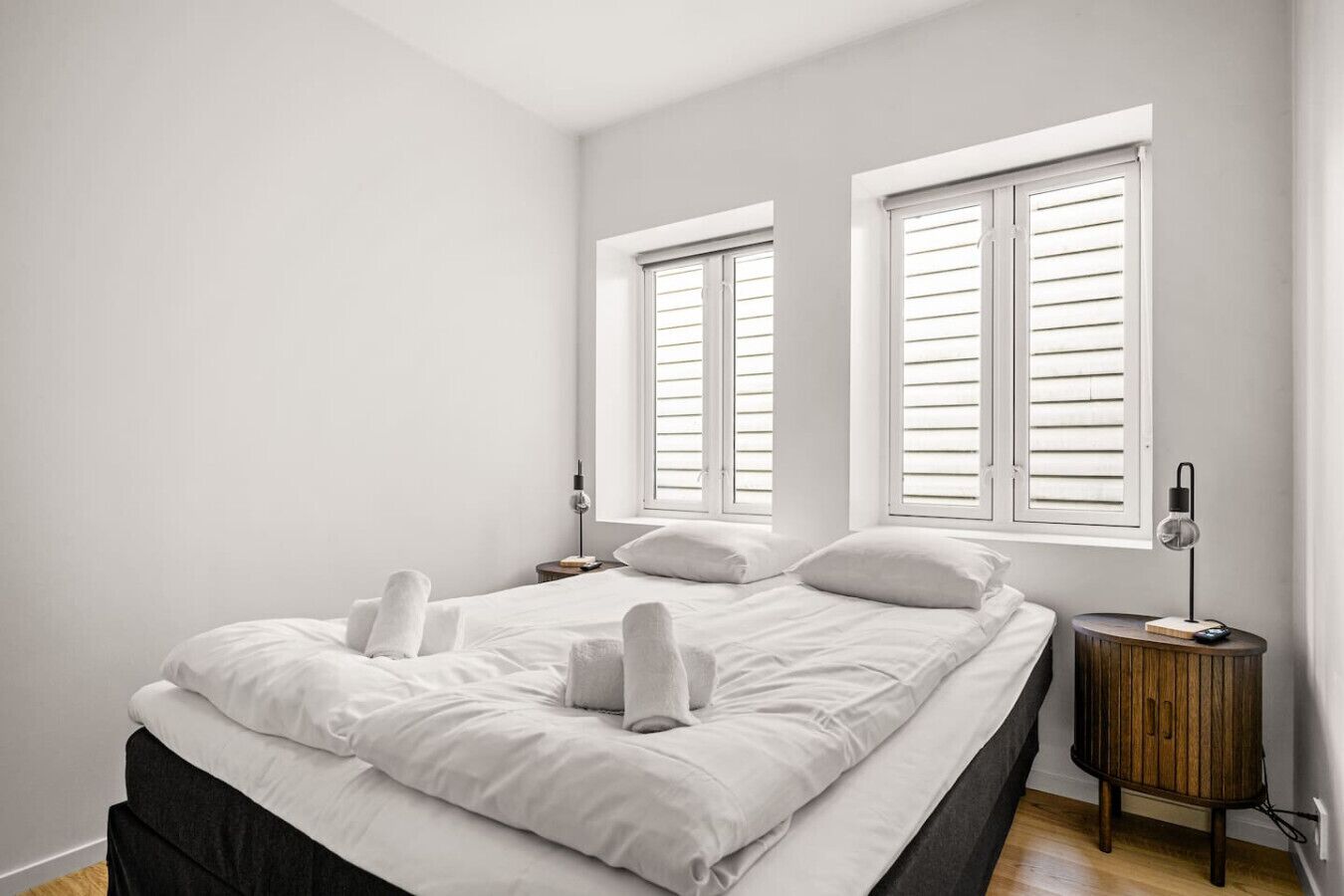 A bedroom with a bed, two bedside tables, and two windows with shutters.